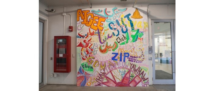 A photo of the foyer leading into Plug In Gallery that shows a large brightly-coloured mural painted with words in a graffiti style. Lanugage is Indigenous slang and Urdu in Arabic script.