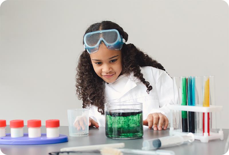A child in a labcoat and goggles looking a beaker with green liquid