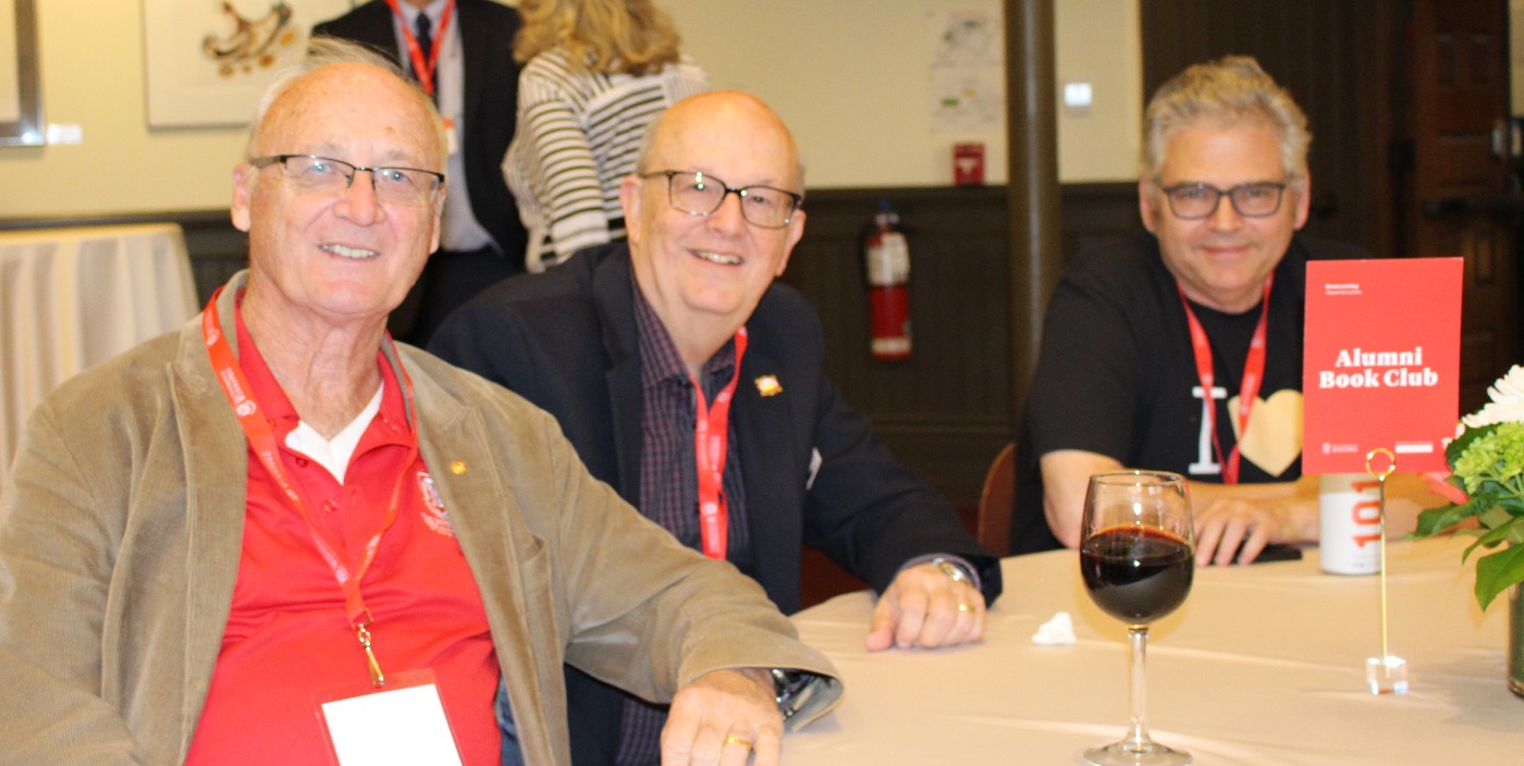 Three men smile at the camera. A red sign on the table reads "Alumni Book Club."