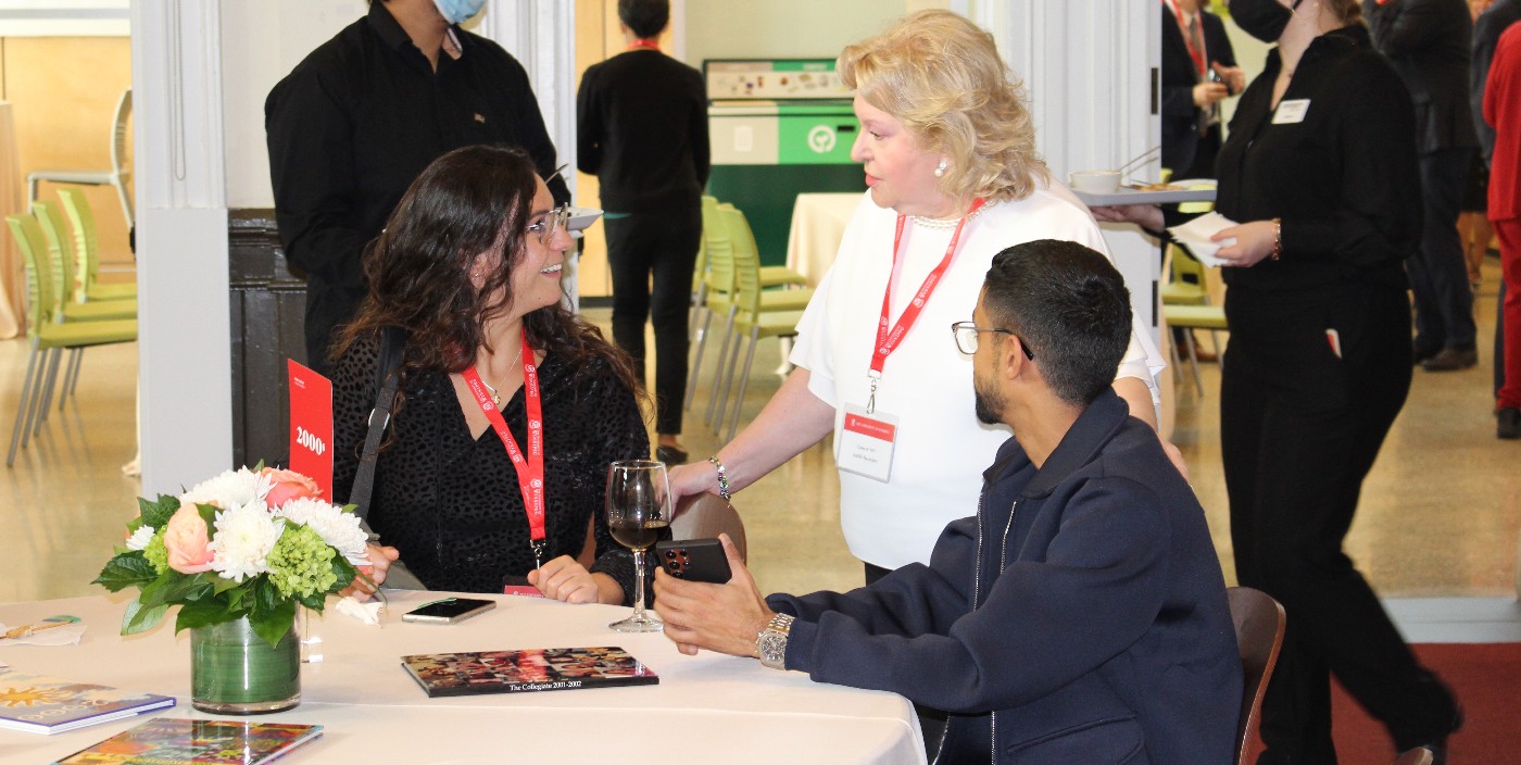 Two people, seated, speak with a woman in a white shirt who is standing tableside.