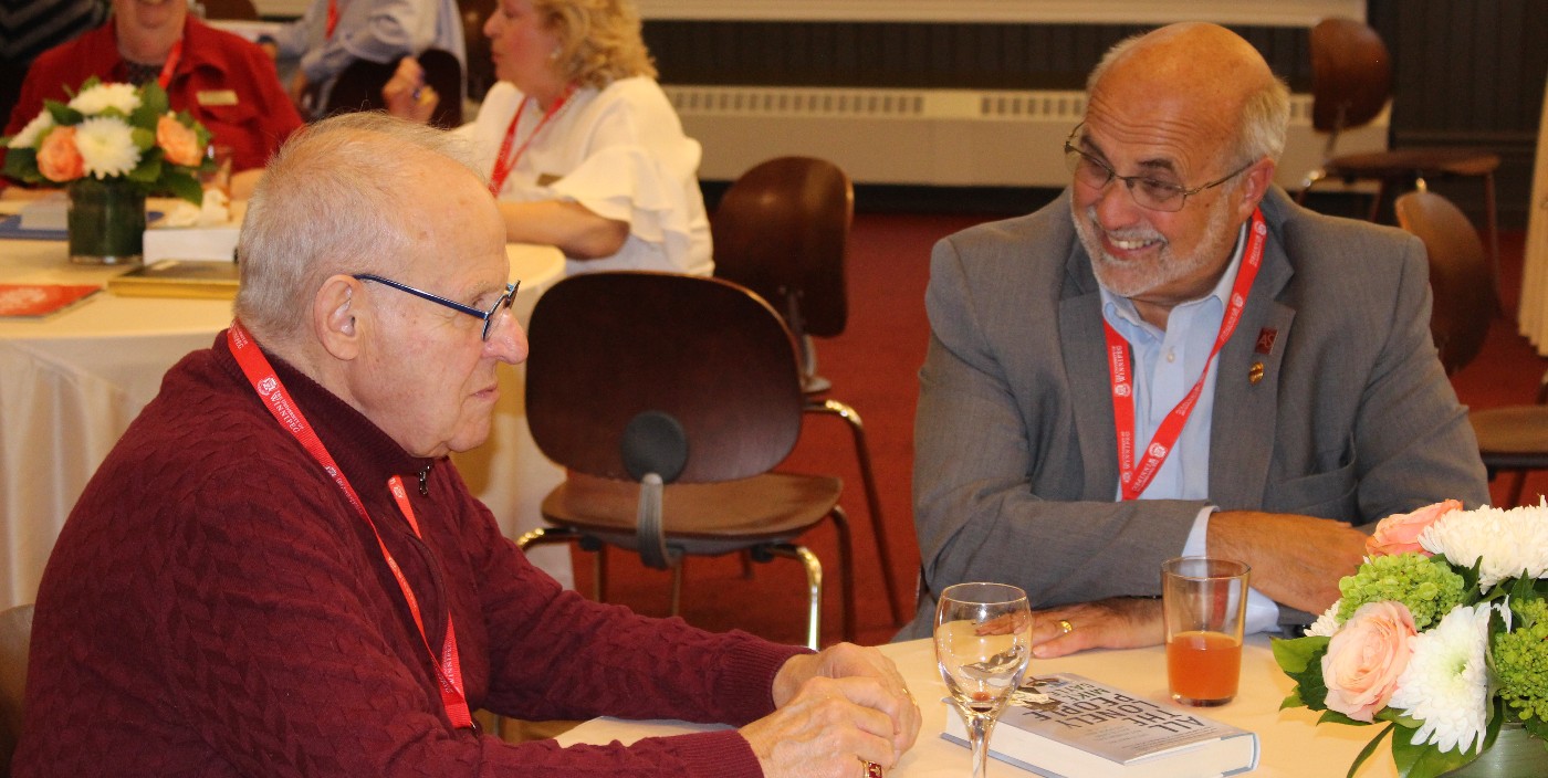 Two men seated at a table speak to each other while seated at a table.