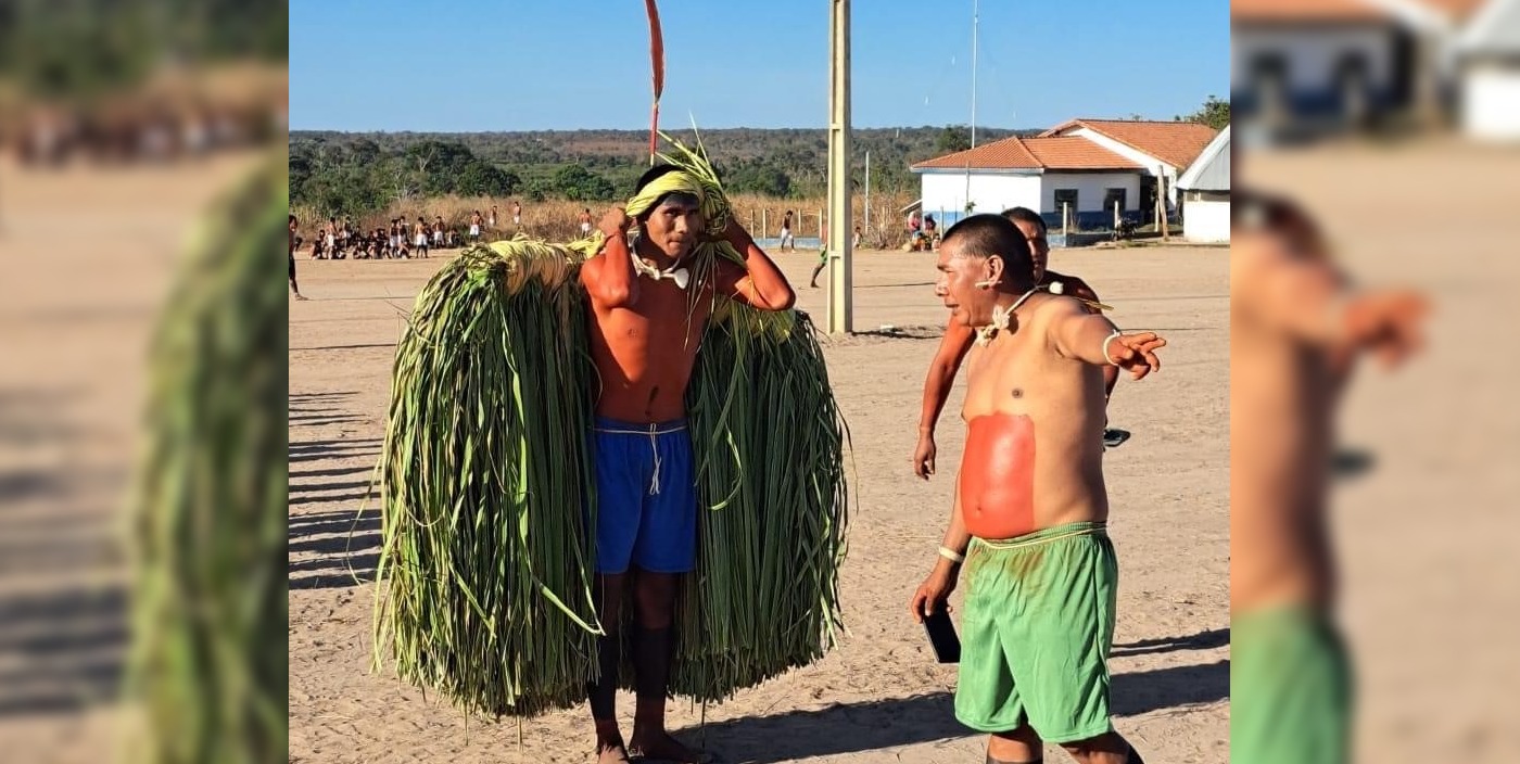 Two Xavante men wear red paint and speak while standing in an open field