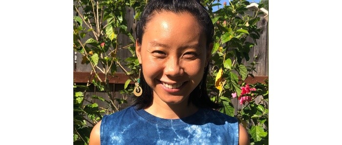 headshot of iris yirei hu facing viewer and standing in front of climbing plant on a trellis. She wears an indigo coloured tie dye t-shirt.