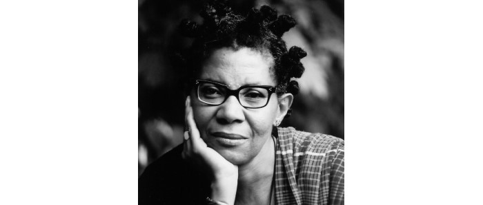 Black & white headshot of M. NourbeSe Philip facing viewer with one hand cupping her cheek. She looks pensively at the viewer with a slight smile. NourbeSe's black hair is knotted in several braids that are tied up on her head. She wears dark rimmed glasses and a plaid top.