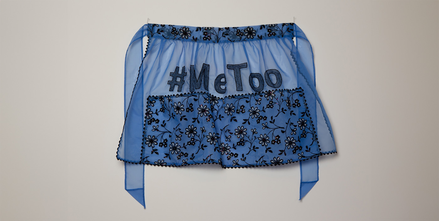 a sheer blue vintage apron hangs on a white wall. It has a black floral pattern along the bottom half and along the waist band. The hashtag #MeToo is embroidered across the middle.
