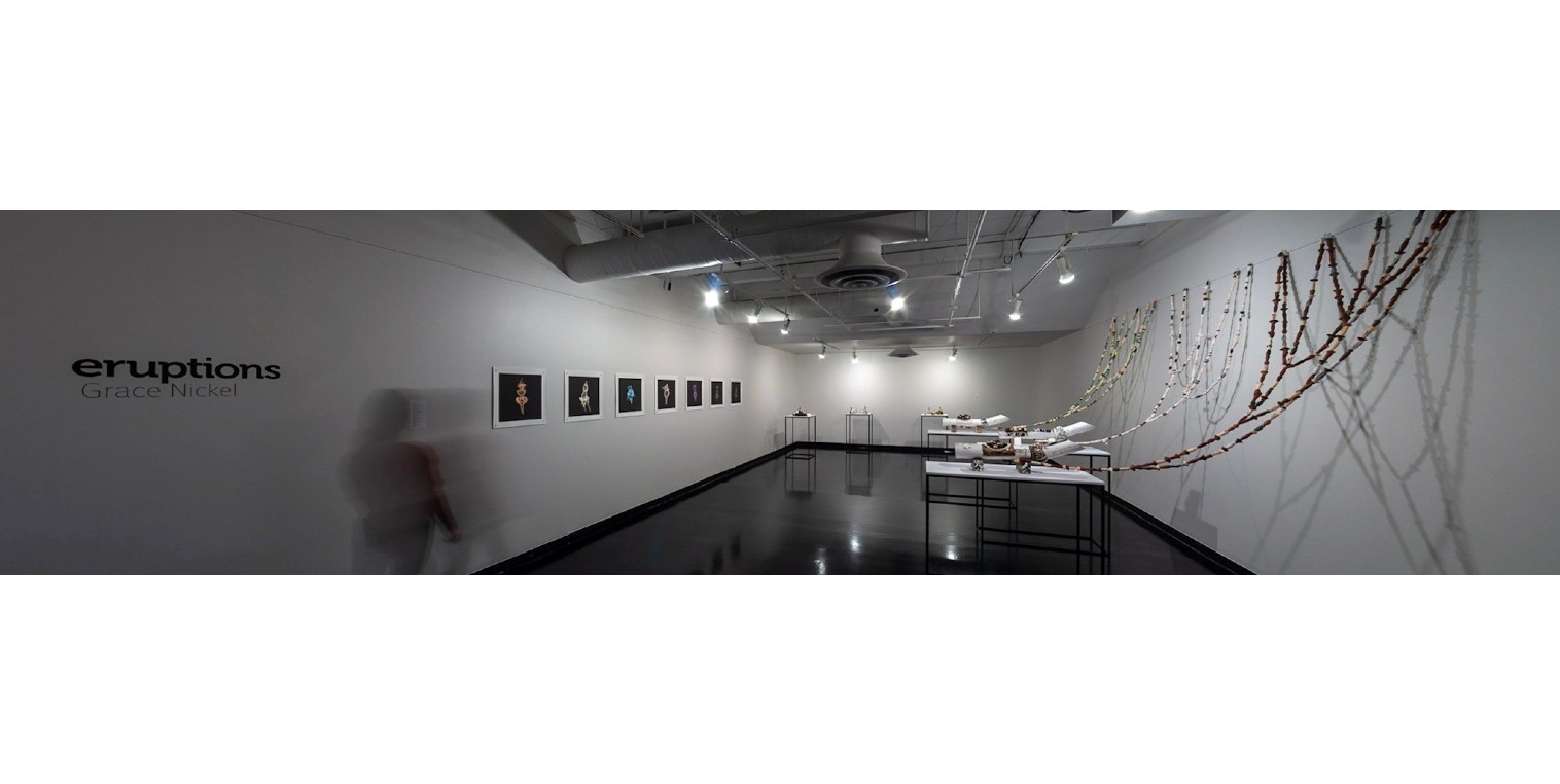 Photo of a blurred person walking in white-walled gallery. There are photos on the left wall and sculptures on the back and right walls.