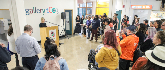 a view in front of the gallery shows a crowd gathered to listen to a person speaking at a podium