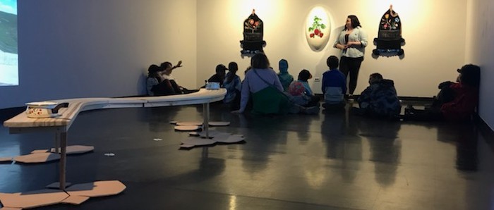 A view of the gallery shows art educator Ruby Bruce standing beside 3 woven and beaded wall hangings on the back wall. She is speaking to a class of children who are seated on the floor in front of her. She looks at a child on the left who has their hand raised to ask a question. In the foreground is a sculpture: a long, low curving wood table with a pot placed atop it at one end and clay tiles beneath it. 