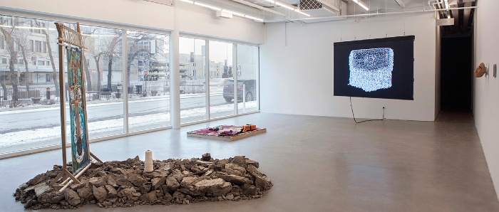 photograph shows view of Sovereign Intimacies exhibit inside Plug In's gallery. Artworks from front to back: sculpture installation of chunks of clay on the ground & vertical painting framed in upright loom; red patterned blanket spread flat & lifted slightly off floor by wood platform; video still of white points of light forming a blanket pattern on a black video screen suspended in the room; two small wood masks & an ipad hung on the wall at right. The left wall is completely windows looking out onto Colony Street in Winnipeg.