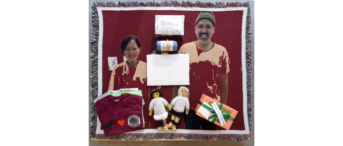 Photo of an installation that shows a red fringed photo blanket of artists Ayumi Goto and Peter Morin standing side by side. On top of the blanket are two handmade dolls dressed in winter clothing and holding hands, a stack of 4 t-shirts and several unwrapped gifts. The blanket lies flat on the floor of the gallery but is raised slightly as it is on top of a low platform.