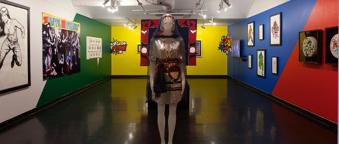 Photo of gallery shows mannequin wearing regalia in foreground in front of red and black button blanket. The gallery walls are painted bright colours, clockwise around the room: white, green, yellow, red and blue. Comic book style graphics that read POW and WOW are on the left and right corners of the back wall. Artworks on wall from left to right: large black & white drawing of a woman superhero in action, large black & white banner of several Indigenous women in a moon-like landscape, a comic speech bubble that has 13 small framed trading cards, a video monitor, 6 drawings in black frames, 2 painted hide drums in black frames.