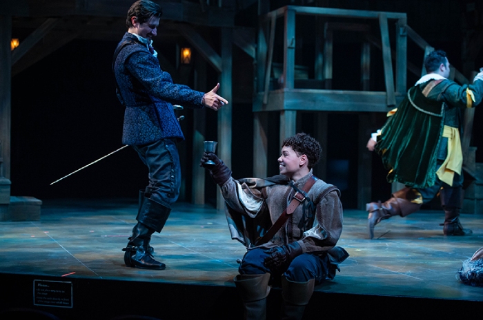 D'Artagnon (Melissa Langdon) sits on the edge of the stage, turned to her right and raising a glass to one of the Muskateers