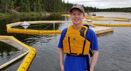 Daniel Denton, wearing a life jacket, stands in front of a lake. In the background, yellow barriers pen in water-testing areas.