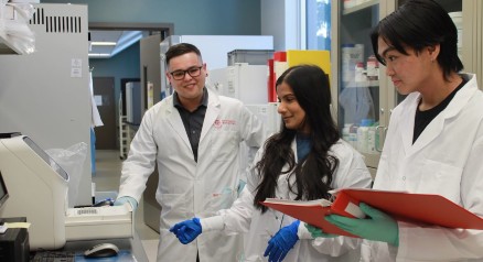 Dr. Sanoji Wijenayake, wearing a lab coat, stands in front of lab equipment. Two fellow researchers stand alongside.