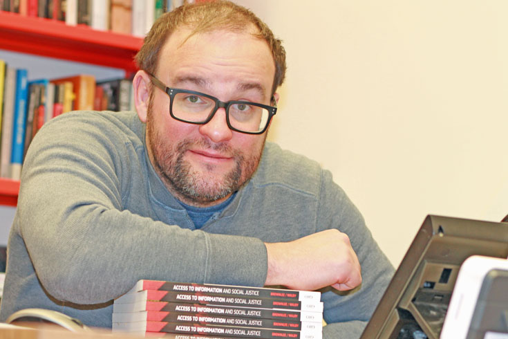 Dr. Kevin Walby, leaning on a stack of books entitled "Access to Information and Social Justice"