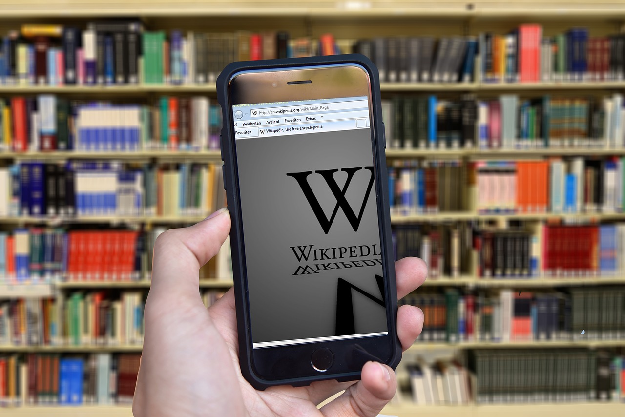 Phone open to Wikipedia page, held up in front of a library stack; Image by Gerd Altmann from Pixabay