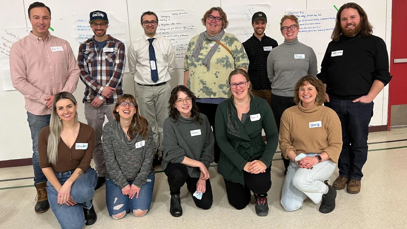 A group of a dozen smiling adults wearing name tags are gathered in front of flip chart paper covered with colourful writing.