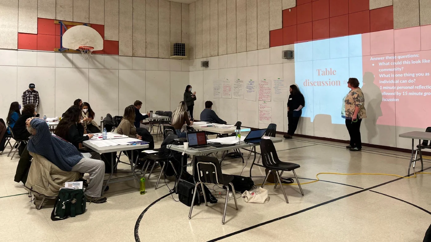 A group of people are gathered in a gymnasium taking part in a community event. Participants sit at long tables with notebooks and other materials engaging with the two presenters, who are leading the discussion using a PowerPoint.