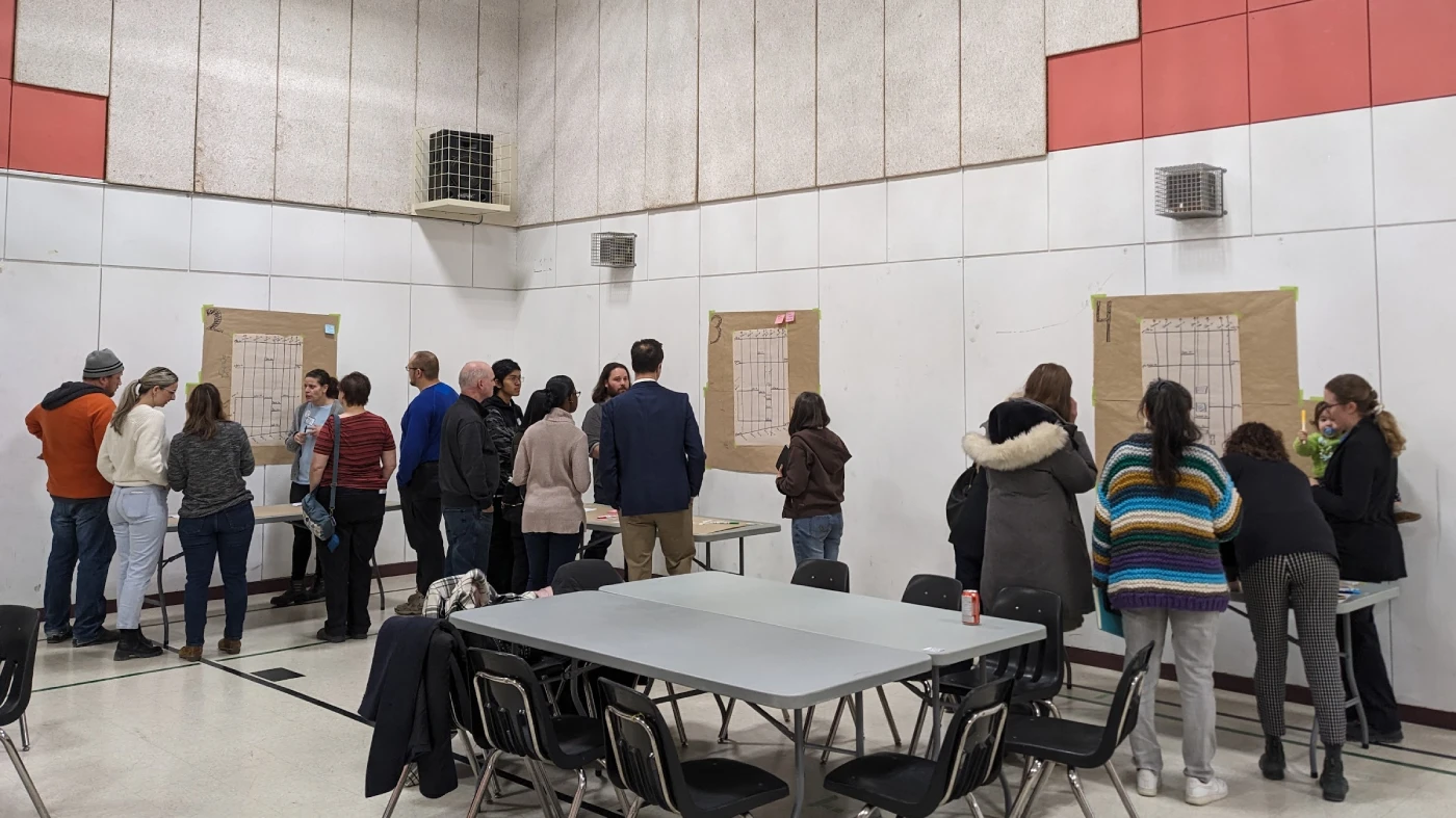 Small groups of people bundled up in sweaters and winter wear are in a community gymnasium. They are clustered around large pieces of paper, collaborating on ideas and jotting them down.