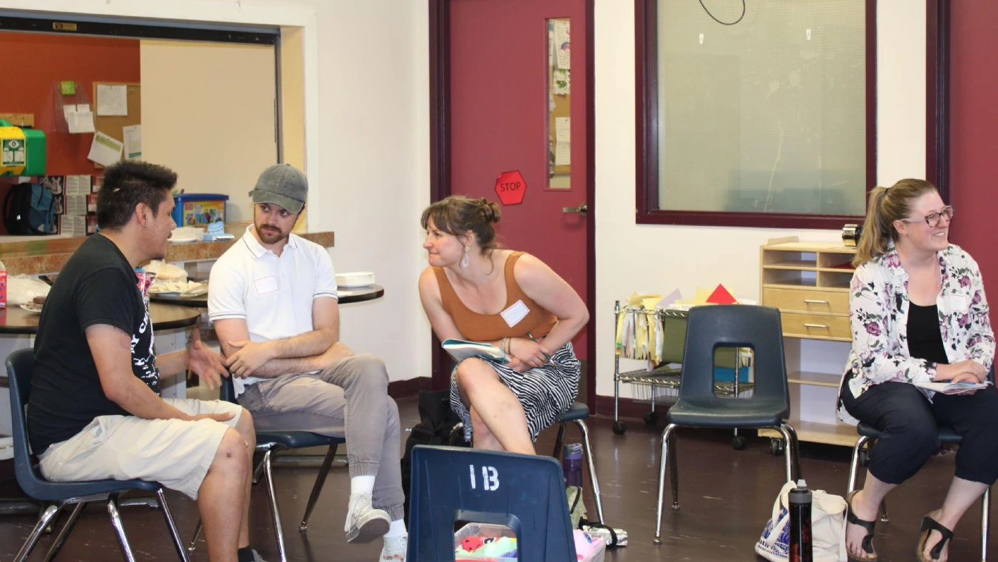 Four adults sit in a semi-circle in a brightly lit community or classroom space with red doors and window trim, round tables, and grey chairs. The group is engaging in a relaxed and attentive manner, and are taking part in a harm reduction workshop.