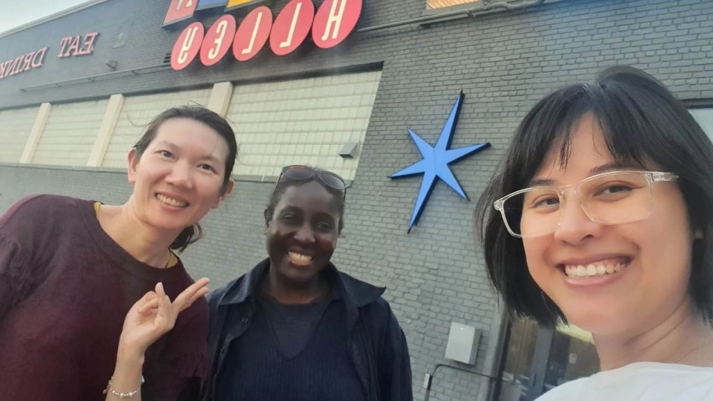 Three people stand around a wooden dining room table surrounded by paper bags. They are adding chips, fruit, and other treats to the bags in preparation for the community scavenger hunt they are planning!