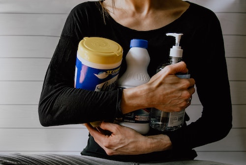 Image of a person holding a bottle of sanitzing wipes, bleach, and hand sanitizer.