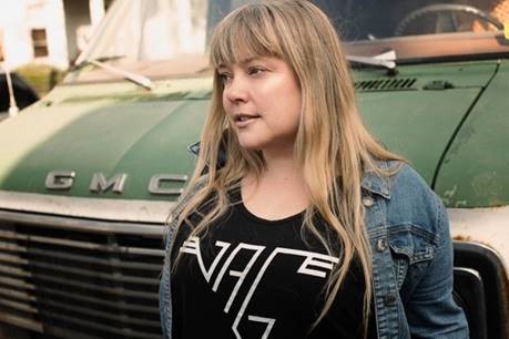Zoe Whittall stands in front of an old, green van wearing a black shirt and denim jacket