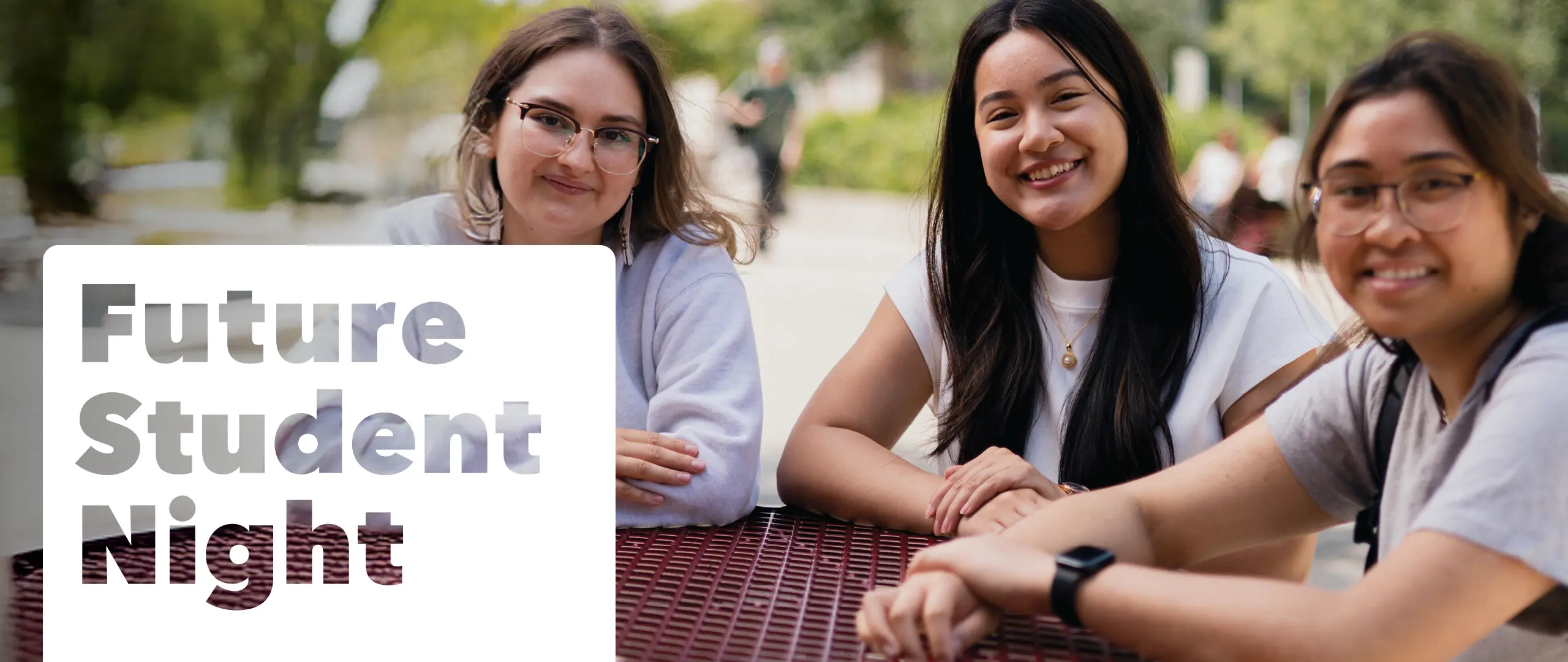Three students sitting at a table outside smiling at the camera the words "Future Student Night" to the left