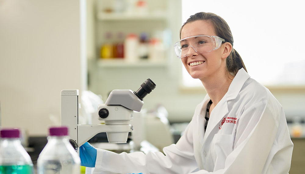 A student in a labcoat with a microscope smiling