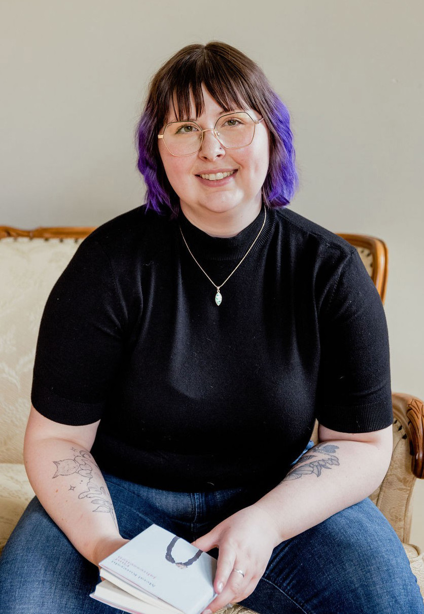 Angela sits on a cream-coloured couch, holding a book in her hands as she smiles at the camera