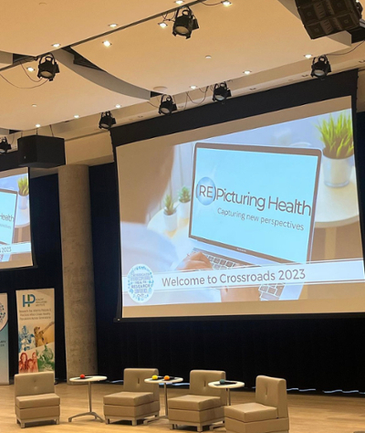 A photo of the conference stage. There are four beige armchairs with side tables between them. A projector screen behind them is displaying an image of a laptop with the screen reading “(Re)Picturing Health: Capturing new perspectives.” A banner across the bottom of the projector screen reads “Welcome to Crossroads 2023.” 