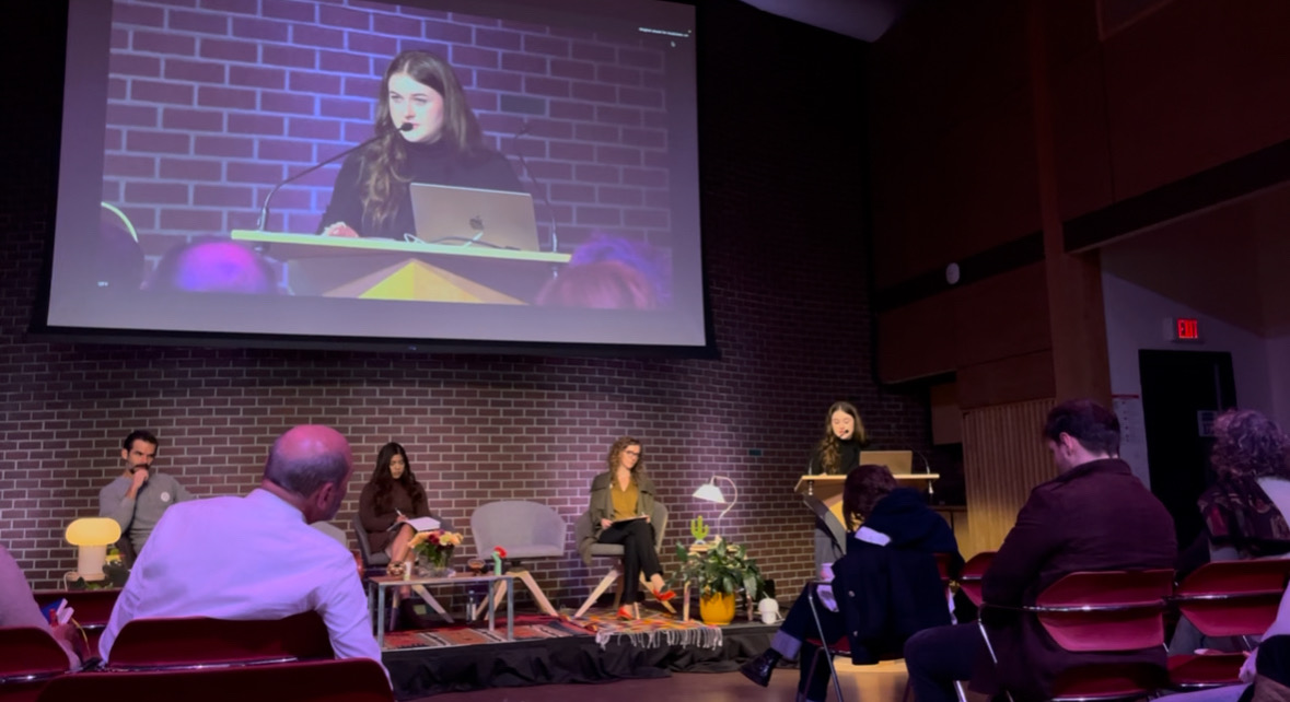 Kenna speaking at a podium while the audience watches