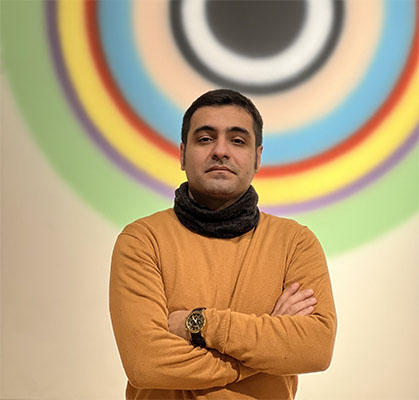  Moeen Didehban, MA in Cultural Studies student, stands in front of a mural of overlapping, colorful circles. His arms are crossed and he is looking at the camera.