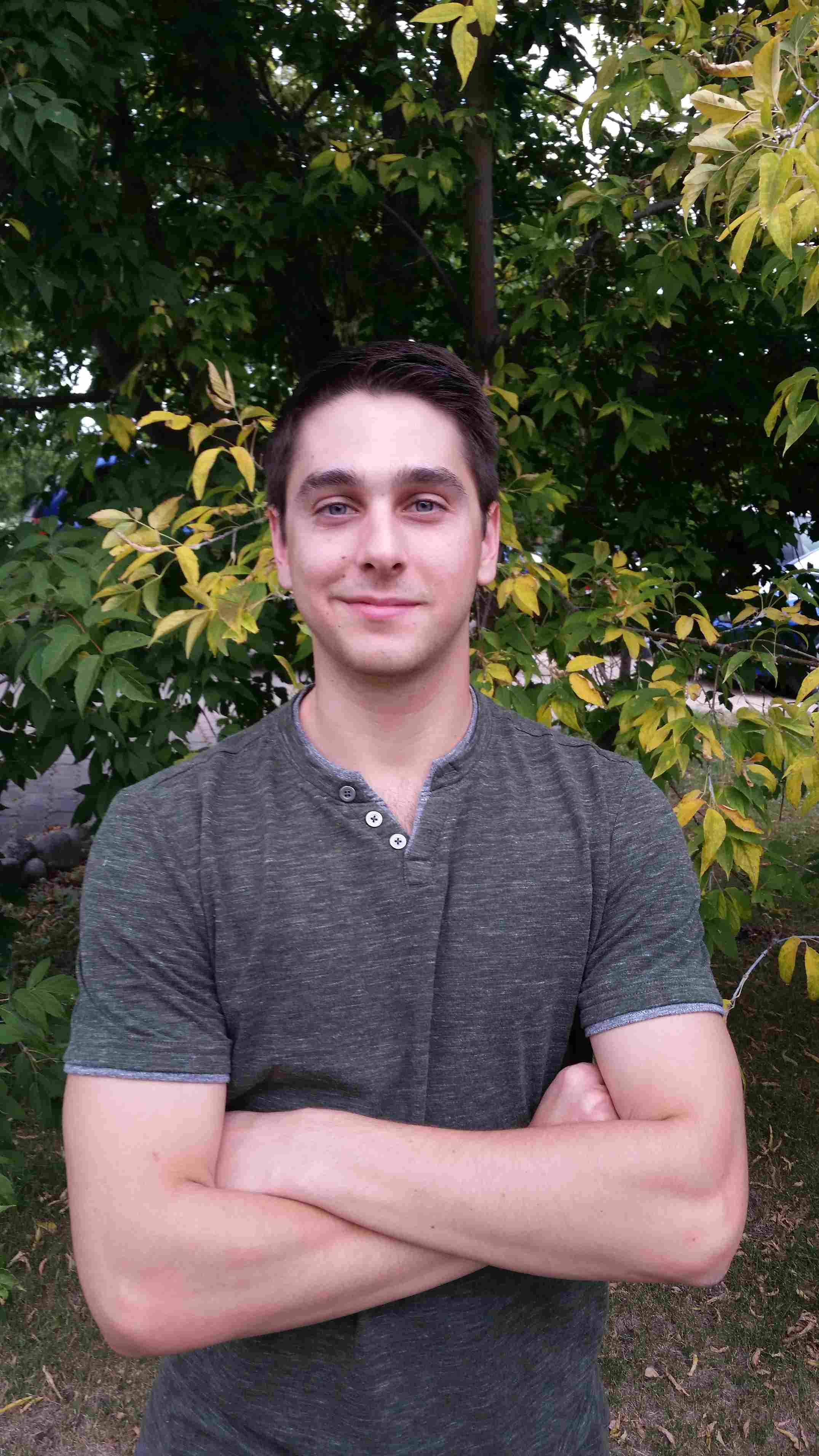 Graduate Student Wr riting Mentor Blake Carter. He is standing in front of a leafy tree, wearing a grey t-shirt. He is smiling and his arms are crossed.