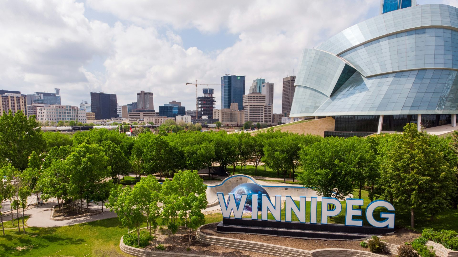An aeriel photo of Winnipeg's The Forks. In the foreground is a large sign spelling out "Winnipeg". It is surrounded by trees. In the middle ground on the right there is the Canadian Museum for Human Rights. In the background is the skyline of downtown Winnpeg. 