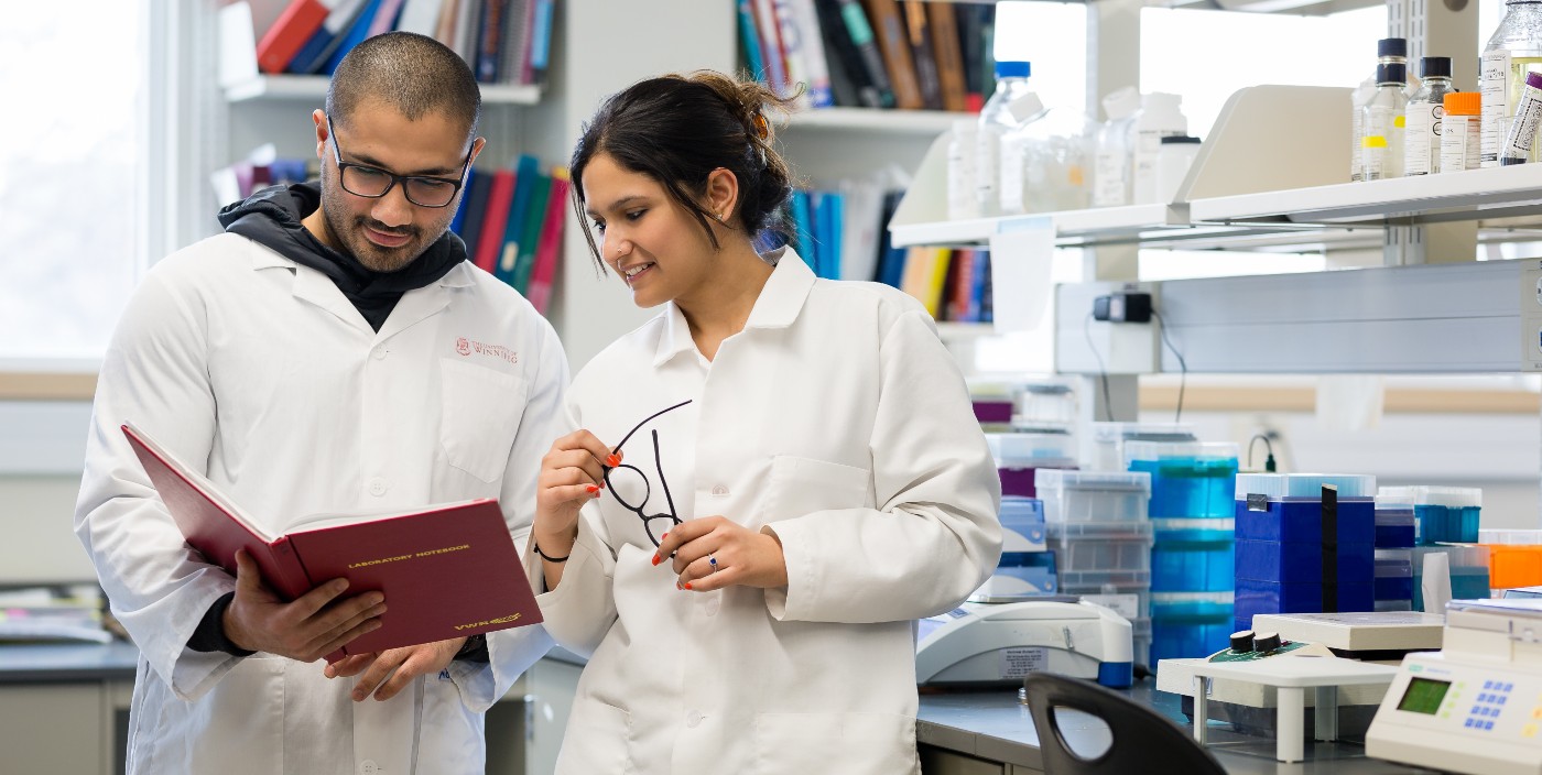 Two students in white lab coats talk while one holds an open folder