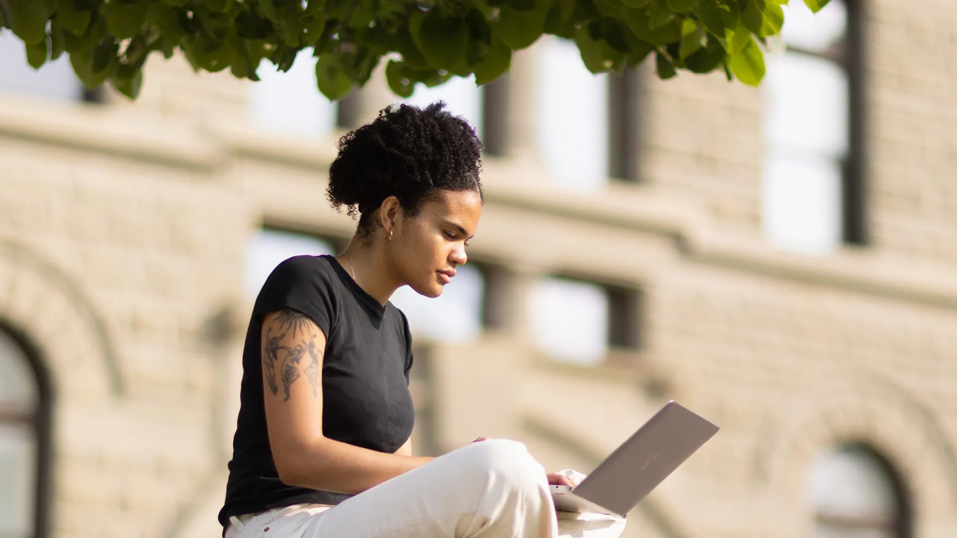 A student is sitting under a tree and working at a laptop computer.