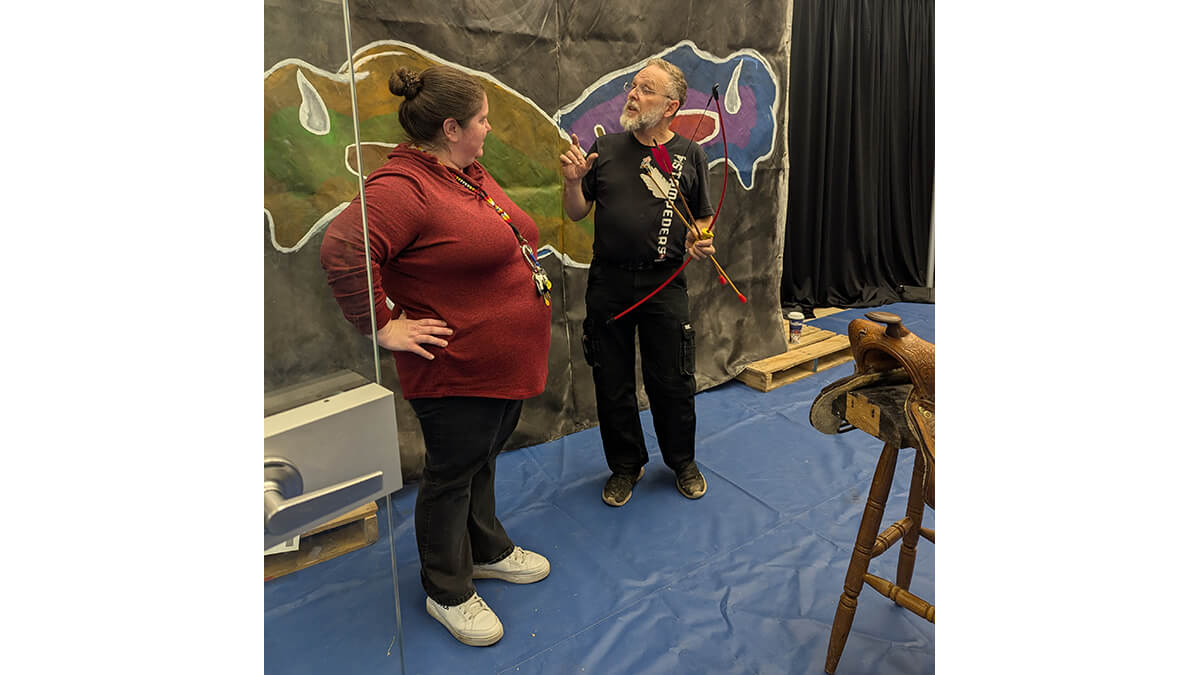 Two people in front of an exhibit one has a bow and arrow, there is a saddle in the foreground