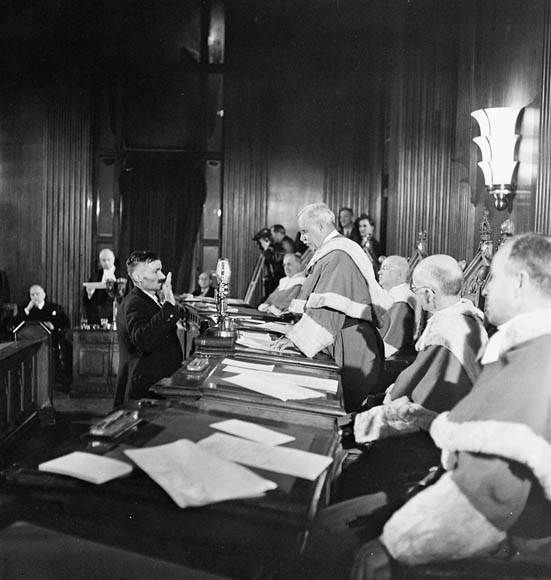 Black of and white image of a man in a courtroom in front of a judge with his hand up taking an oath of allegiance