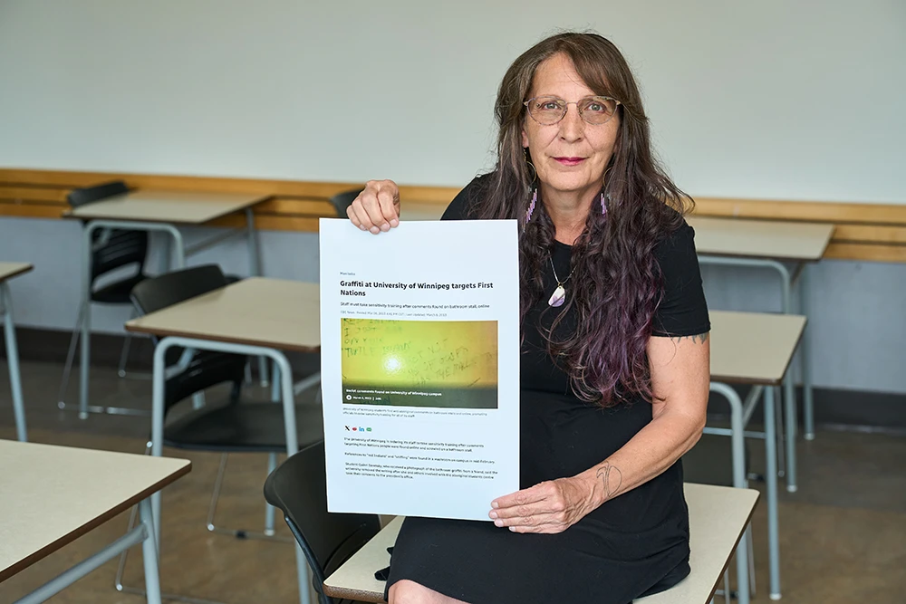 Karen Froman in a classroom sitting on a desk, smiling and holding a document