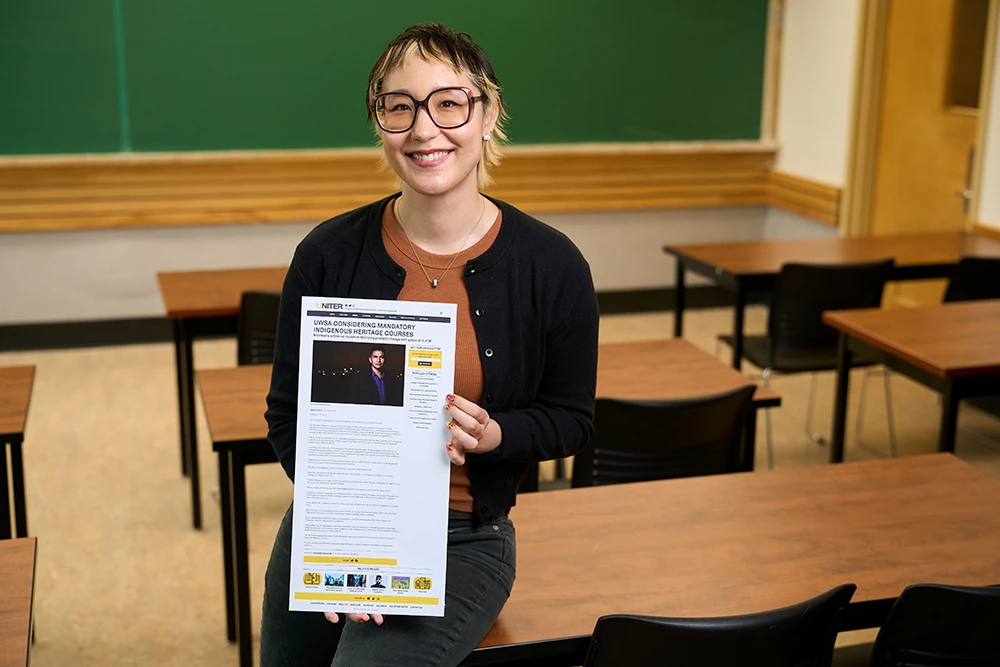 Leah Kuragano in a classroom sitting on a desk, smiling and holding a copy of a newspaper article.