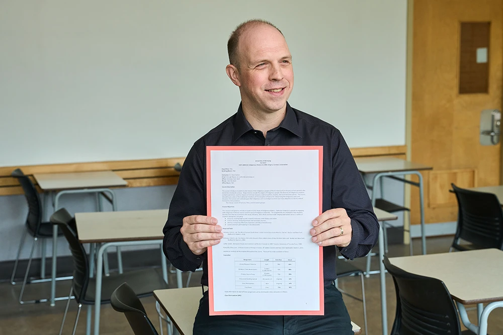 Ryan Eyford in a classroom sitting on a desk, smiling and holding a document