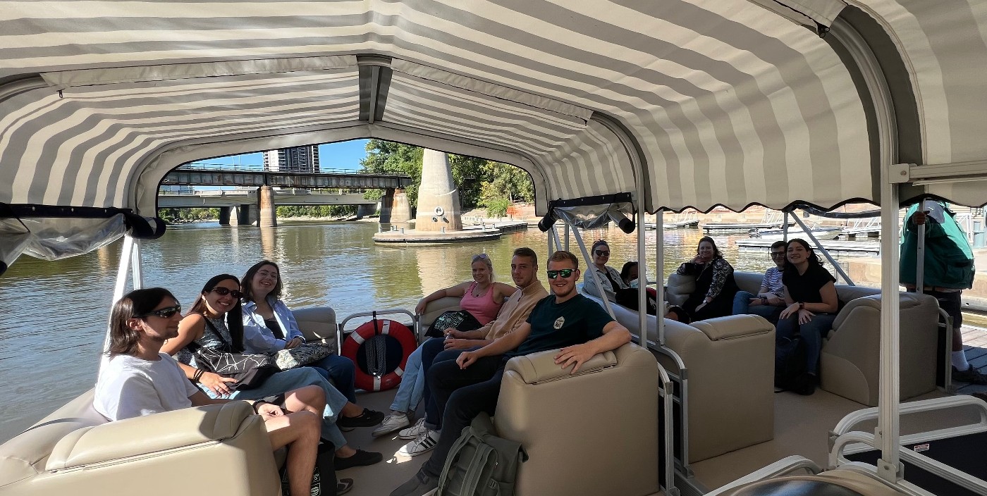 Students sit, shaded, under a canopy on a pontoon boat. The boat is on the Red River, about to take the students on a tour.
