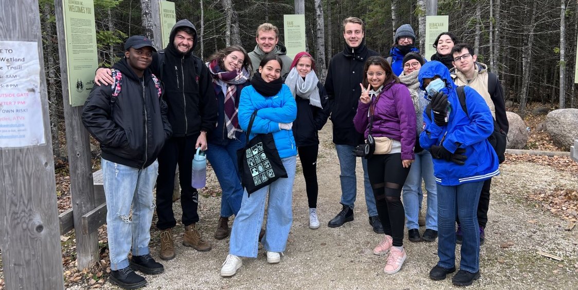 Several students smile to camera at the entrance of a trail. Large trees can be seen in the background.