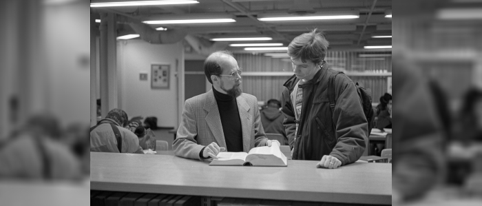 Black and white photo of two people standing at counter and looking through book
