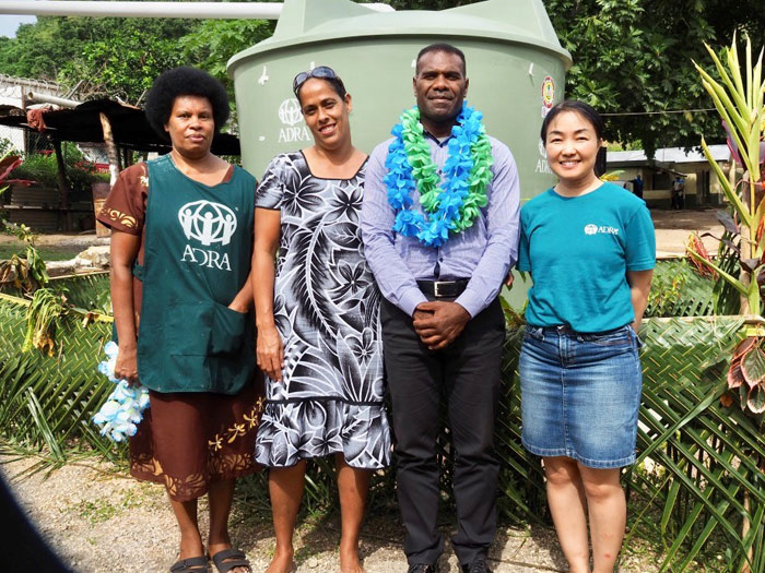 Titima (R) with ADRA staff in front of a water tank for the low Risk Detention Center