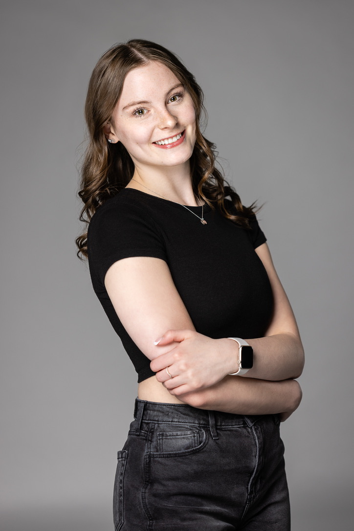 A white woman with long brown curly hair, wearing a black short sleeved shiny and black jeans. 