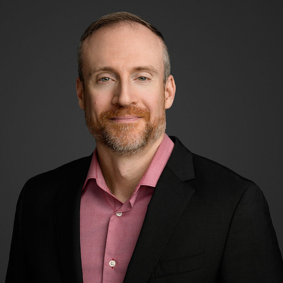A man with a reddish beard and short hair, wearing a black blazer and red/white small striped shirt. Photo credit: Gregory Kennedy.