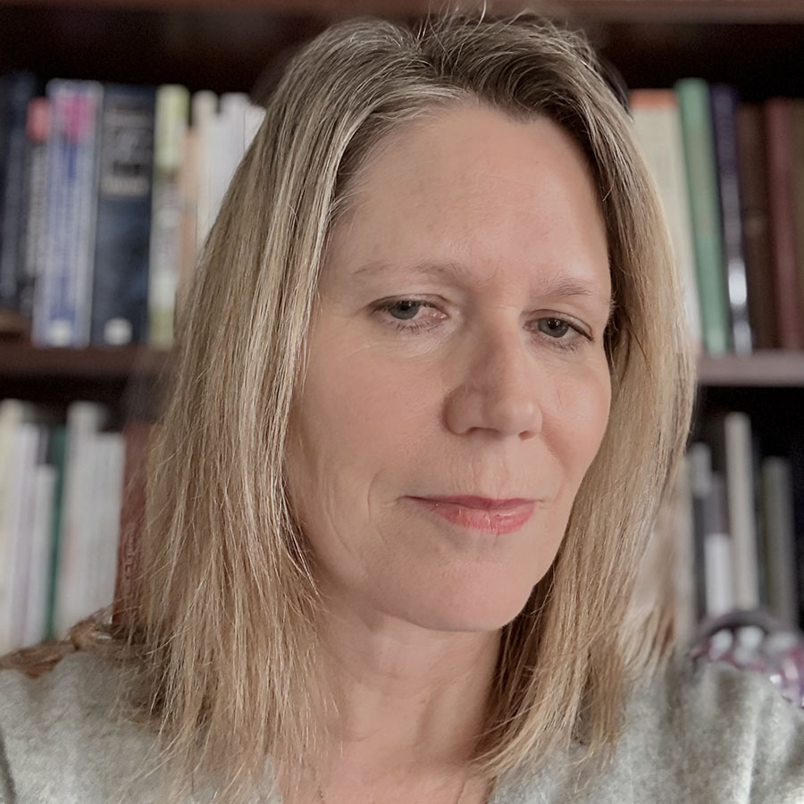 A woman with shoulder length blonde hair, wearing a grey sweater, sitting in front of a bookcase. Photo credit: Marie Carrière.