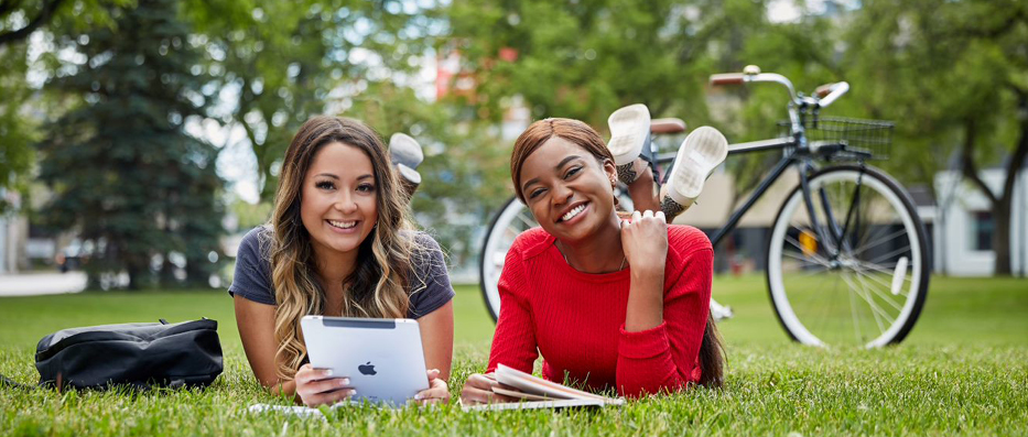 Two women students with ipad and notebook, lying on front lawn